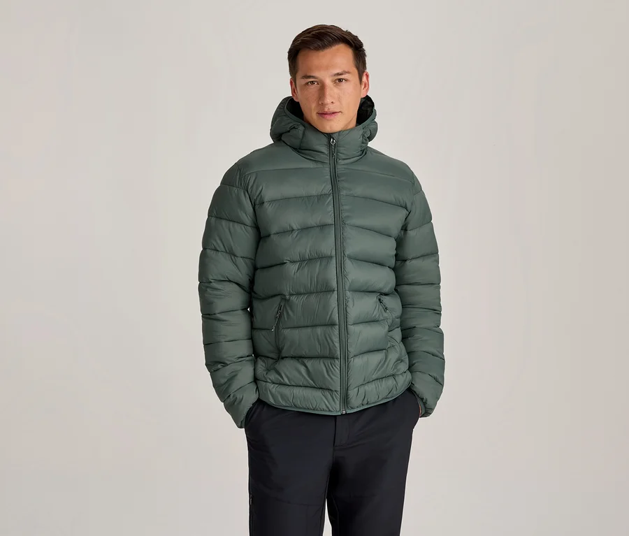 Homme portant une doudoune verte à capuche et un pantalon foncé, debout les mains dans les poches.