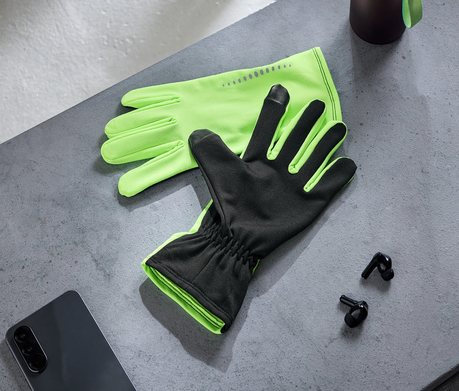 Une paire de gants vert fluo et noirs avec des bouts de doigts tactiles repose sur une surface grise à côté d'un smartphone foncé et de deux écouteurs sans fil noirs. Une bouteille sombre est partiellement visible.