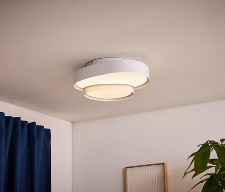 Un plafonnier circulaire blanc et superposé est allumé sur un plafond blanc. À gauche, un rideau bleu foncé est suspendu, et à droite, une plante verte et un tableau abstrait encadré.