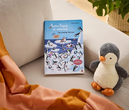 Sur un canapé, on trouve le livre pour enfants "Gustav Pinguin ist unterwegs Eine wimmelige Weltreise" (Exklusive Sonderausgabe), une peluche pingouin et une couverture rayée orange et pêche.