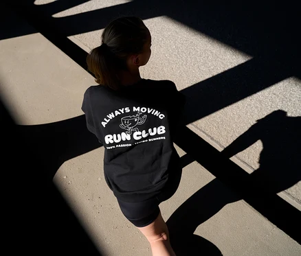 Femme de dos portant un sweatshirt noir avec l'imprimé « Always Moving Run Club » et des leggings de sport courts « ActivePower » anthracite sur un chemin ombragé.