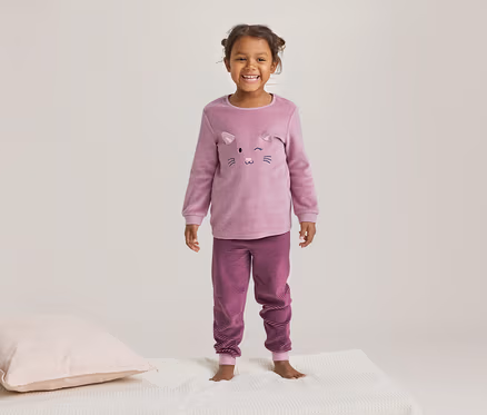 Petite fille souriante en pyjama rose à motif de chat et pantalon rayé, debout sur une couverture blanche à côté d'un oreiller rose.