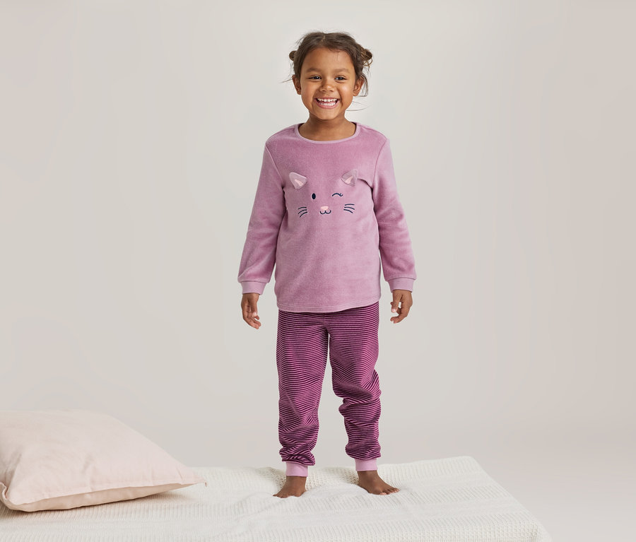 Petite fille souriante en pyjama rose à motif de chat et pantalon rayé, debout sur une couverture blanche à côté d'un oreiller rose.