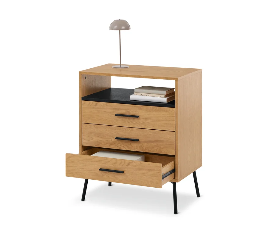Commode en bois clair avec trois tiroirs, une étagère ouverte contenant des livres et une lampe grise sur le dessus. Le tiroir du bas est ouvert, révélant un objet blanc.