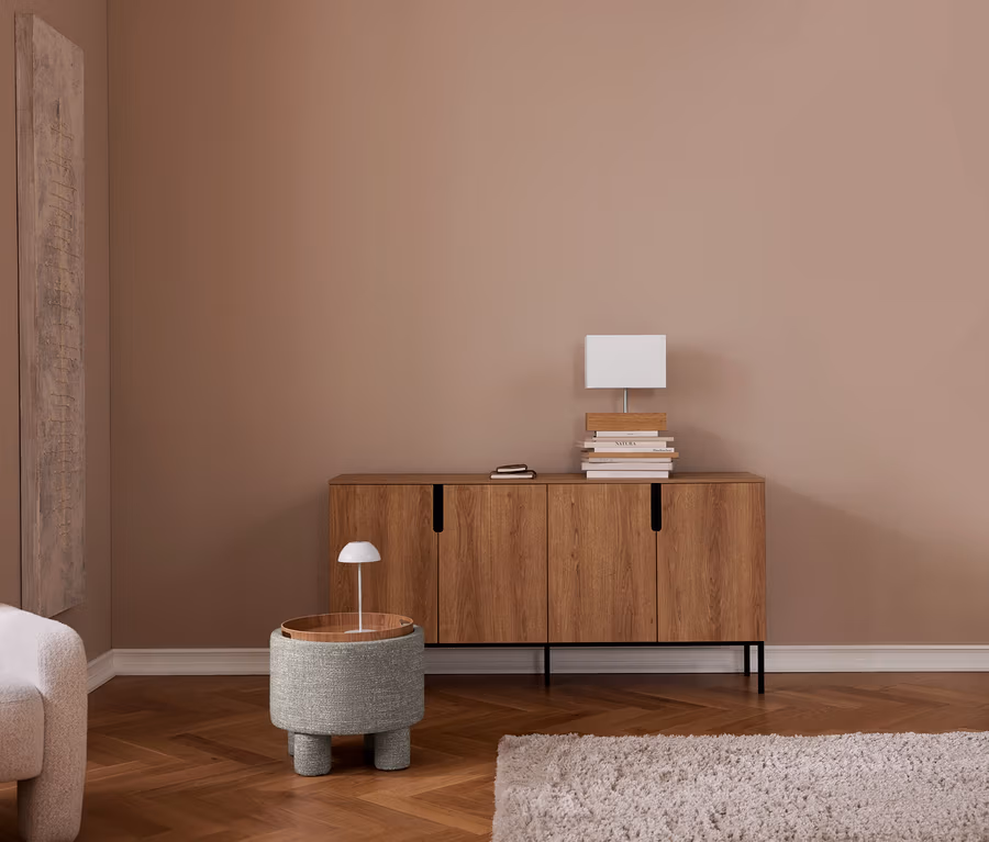 Un salon aux murs couleur taupe et parquet à chevrons. À gauche, un fauteuil beige et une haute œuvre d'art murale. À droite, un buffet en bois avec des livres et une lampe blanche. Devant, un tabouret rond rembourré avec un plateau en bois et une petite lampe. Un tapis clair est au sol.