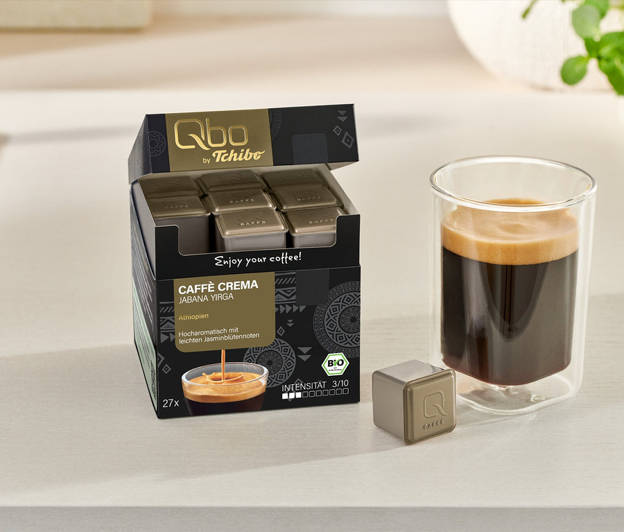 Boîte ouverte de Qbo Caffè Jabana Yirga avec des capsules, une tasse de café et une capsule individuelle sur une surface claire.