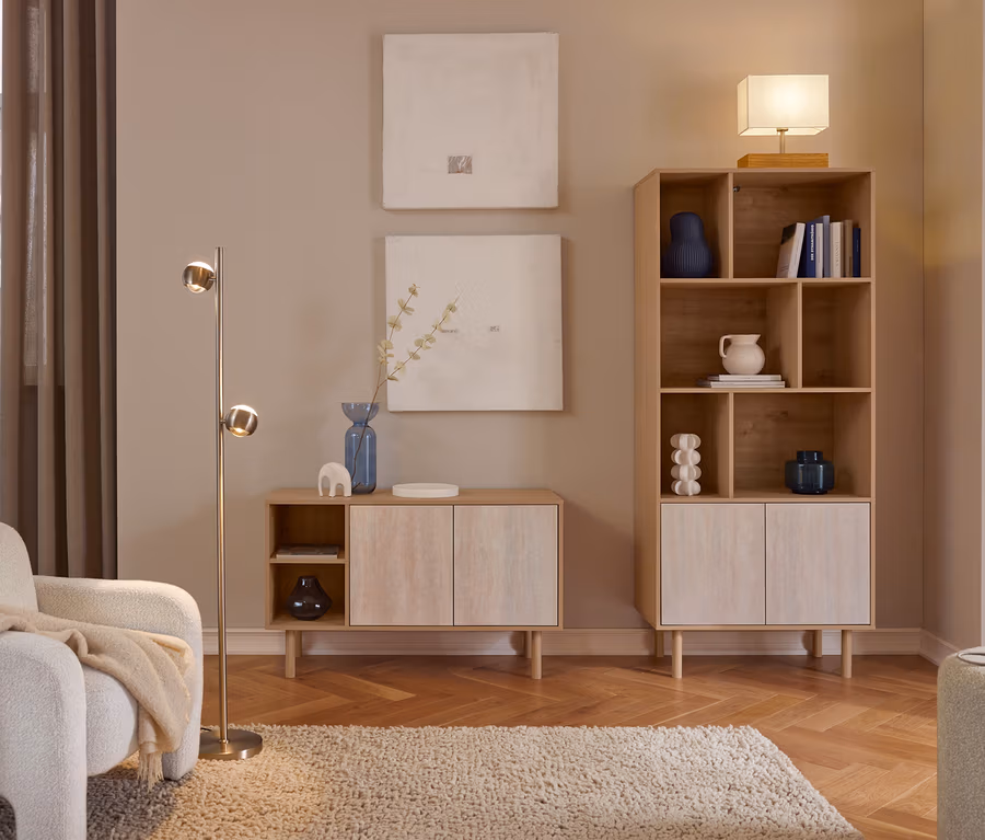 Un salon confortable avec des meubles en bois clair. Un fauteuil blanc avec un plaid beige est placé à côté d'un lampadaire en laiton. Deux tableaux abstraits sont accrochés au-dessus d'un meuble bas avec un vase bleu. Une haute bibliothèque contient des objets de décoration et des livres, surmontée d'une lampe de table. Un tapis shaggy recouvre un parquet à chevrons.