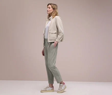 Femme de profil portant une veste beige, un haut blanc et un pantalon de jogging Street One vert clair, une main dans la poche. Elle porte des baskets beiges et blanches.