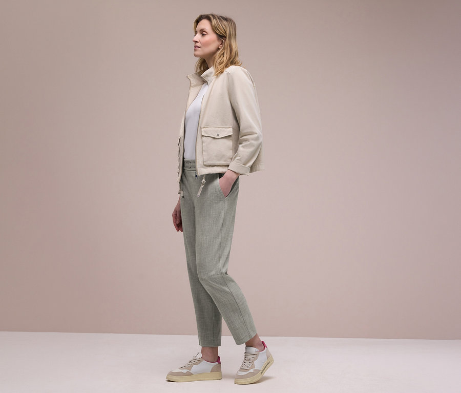 Femme de profil portant une veste beige, un haut blanc et un pantalon de jogging Street One vert clair, une main dans la poche. Elle porte des baskets beiges et blanches.