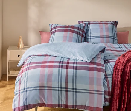 Un lit avec une housse de couette et des oreillers à carreaux bleus, roses et rouges. Un plaid rouge est drapé sur le côté droit. Une table de chevet avec une lampe est à gauche.