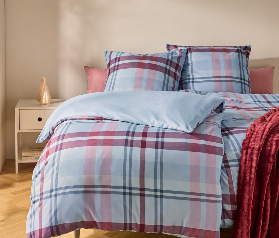 Un lit avec une housse de couette et des oreillers à carreaux bleus, roses et rouges. Un plaid rouge est drapé sur le côté droit. Une table de chevet avec une lampe est à gauche.