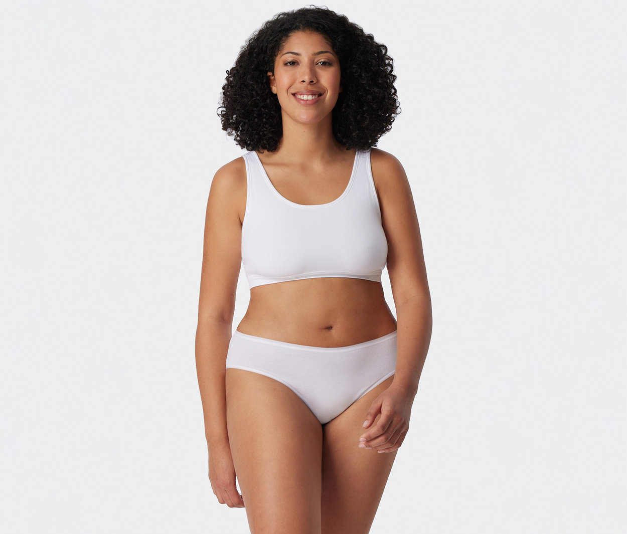Femme souriante aux cheveux bouclés portant un soutien-gorge de sport blanc et une culotte blanche.