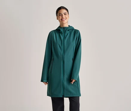 Une femme souriante portant un manteau long softshell vert émeraude à capuche et un pantalon noir sur un fond gris clair.