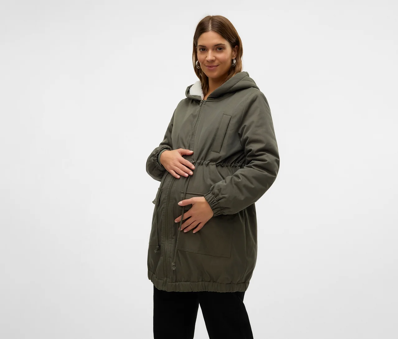 Femme enceinte portant une parka verte avec une capuche doublée de fausse fourrure blanche et un cordon de serrage à la taille, tenant son ventre. Elle porte également un pantalon noir.