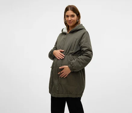 Femme enceinte portant une parka verte avec une capuche doublée de fausse fourrure blanche et un cordon de serrage à la taille, tenant son ventre. Elle porte également un pantalon noir.