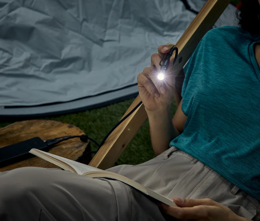 Femme assise dans une tente, lisant un livre avec une petite lampe de poche qu'elle tient.