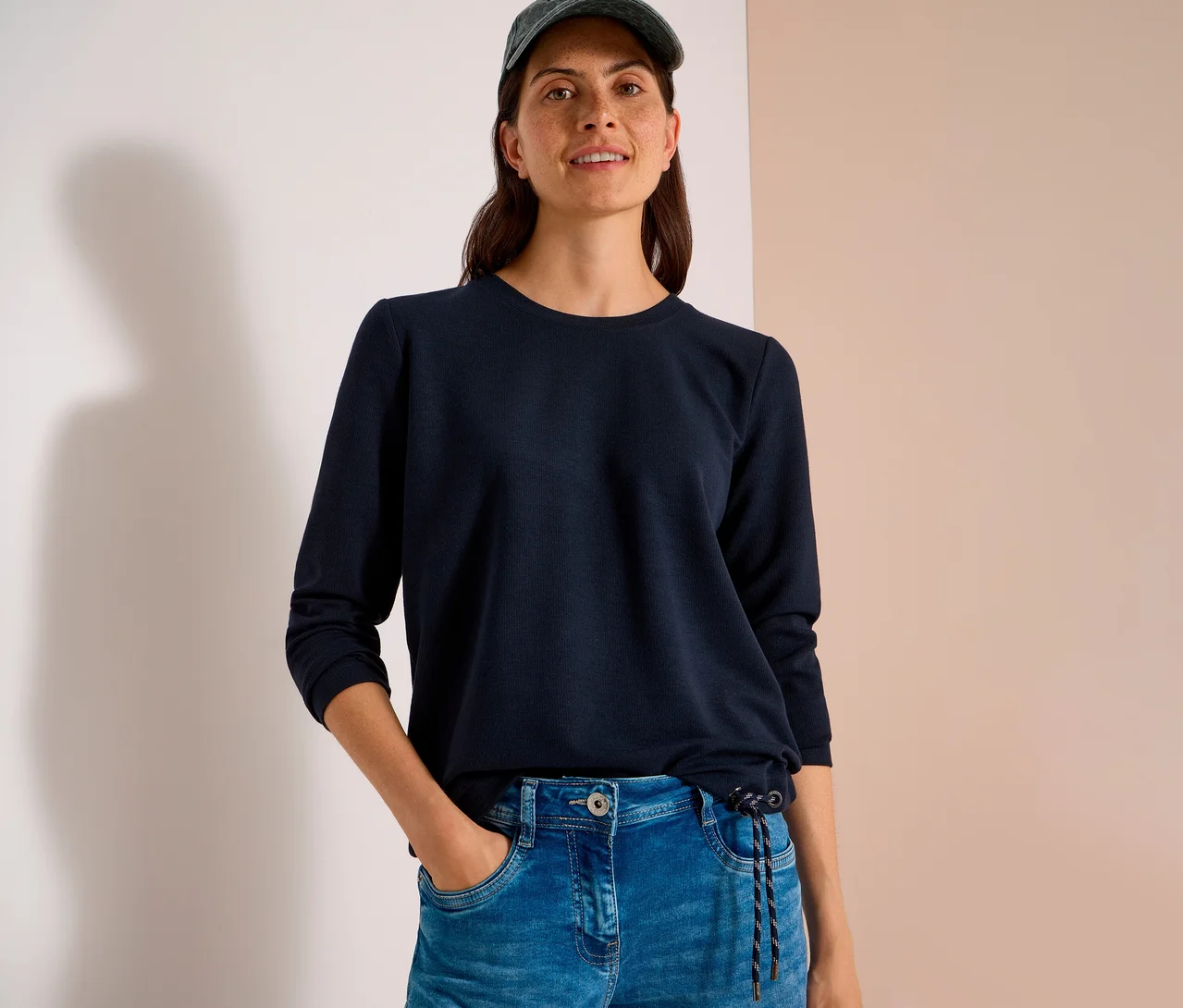 Une femme souriante porte un t-shirt CECIL bleu foncé à manches longues avec cordon de serrage, un jean et une casquette. Sa main est dans sa poche.