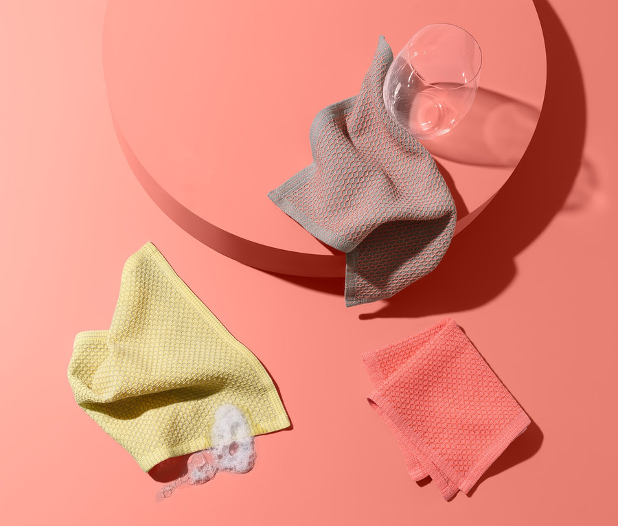 Trois lavettes sur un fond couleur saumon : une jaune avec des restes de liquide vaisselle, une rose et une grise sur un socle rose à côté d'un verre.