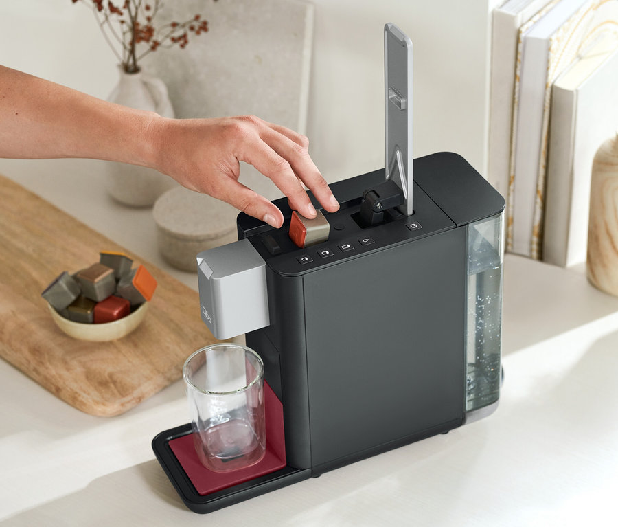 Une main appuie sur un bouton d'une machine à café Qbo ESSENTIAL Dark Berry. À côté, il y a un verre et un bol de capsules.