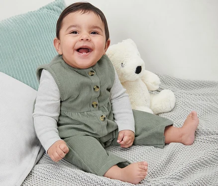 Un bébé souriant est assis sur une couverture tricotée grise à côté d'un ours en peluche et porte une salopette en velours.