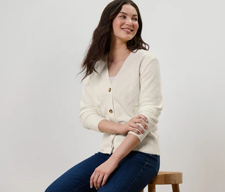 Une femme est assise sur un tabouret et porte un cardigan blanc crème à structure tricotée.