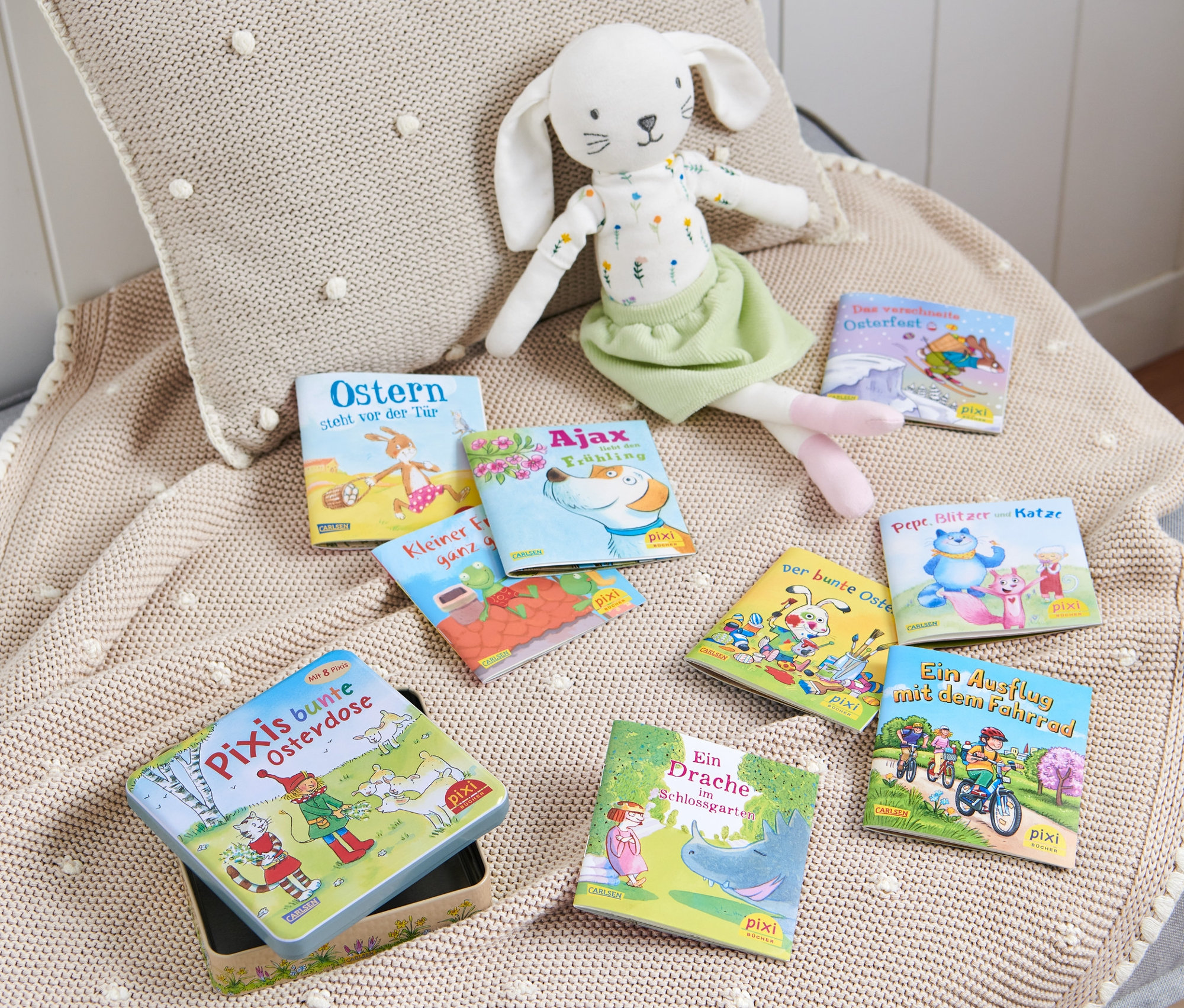 Sur une couverture en tricot beige se trouvent une boîte en métal ouverte avec un livre Pixi et d'autres livres Pixi, un lapin en peluche et un coussin.