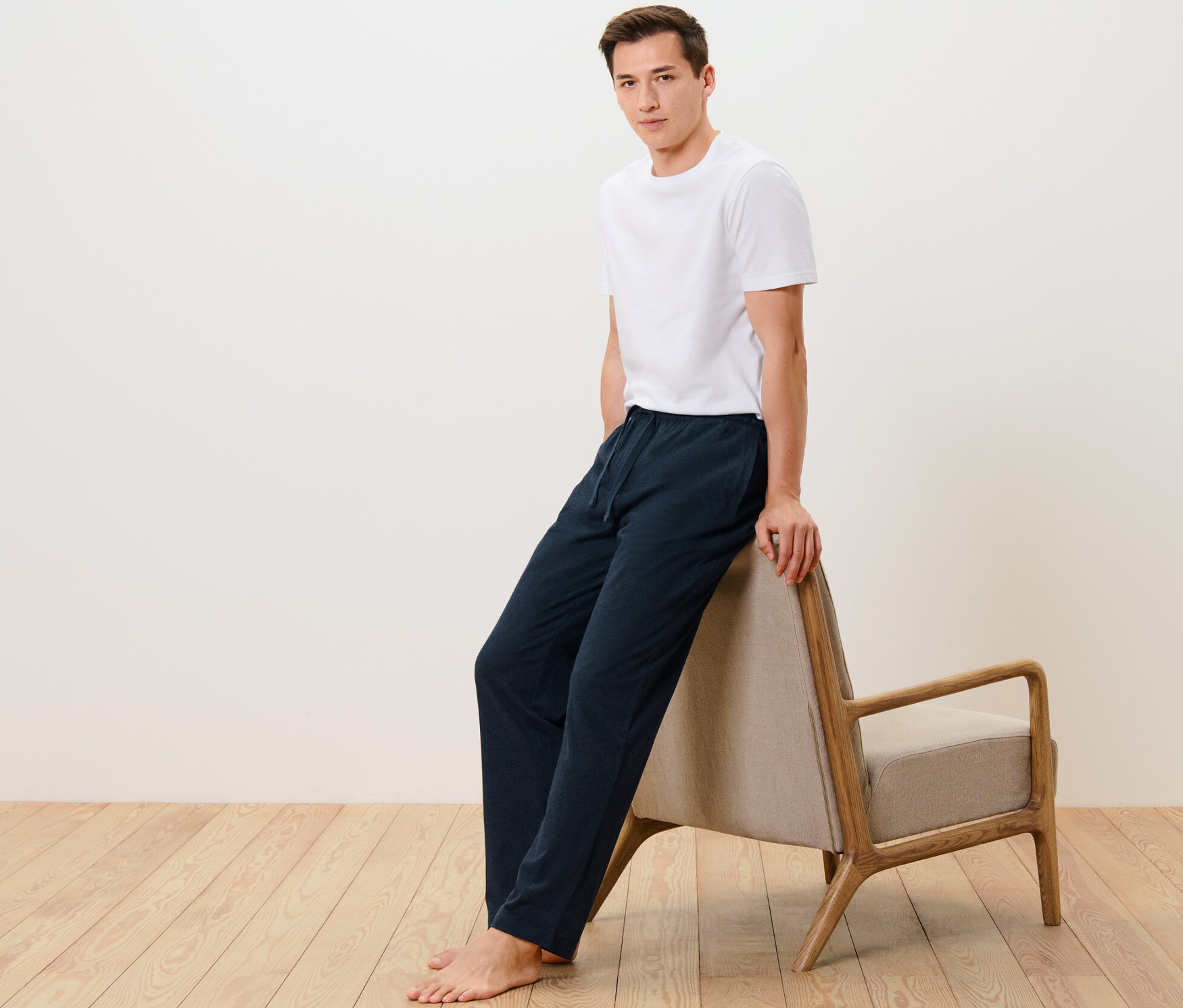 Homme en t-shirt blanc et pantalon relax appuyé sur un fauteuil beige.