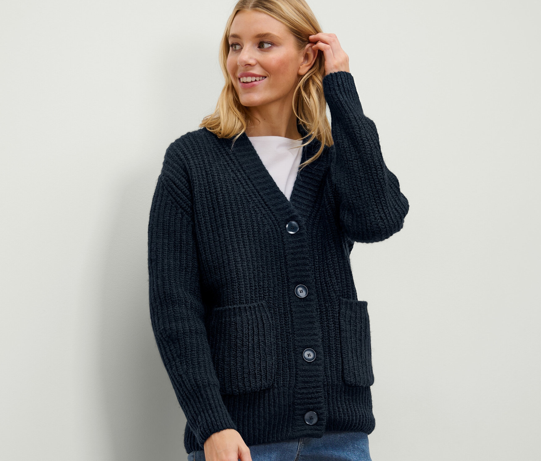 Une femme blonde porte un cardigan en grosse maille côtelée bleu foncé et touche ses cheveux.