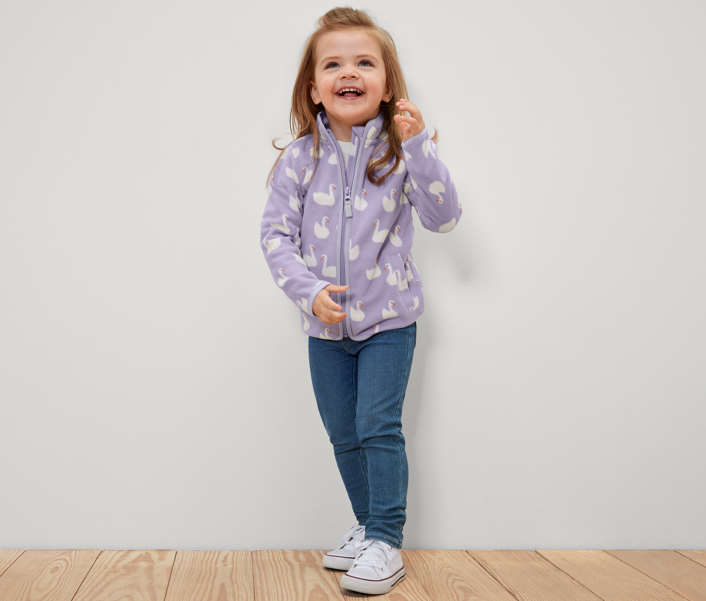 Fillette souriante portant une veste polaire pour enfant à motif cygne, un jean et des baskets.
