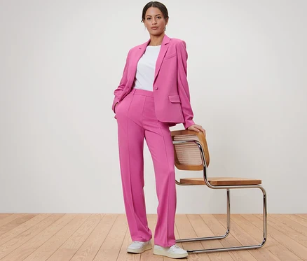 Femme posant en blazer et pantalon en jersey rose à côté d'une chaise en bois.