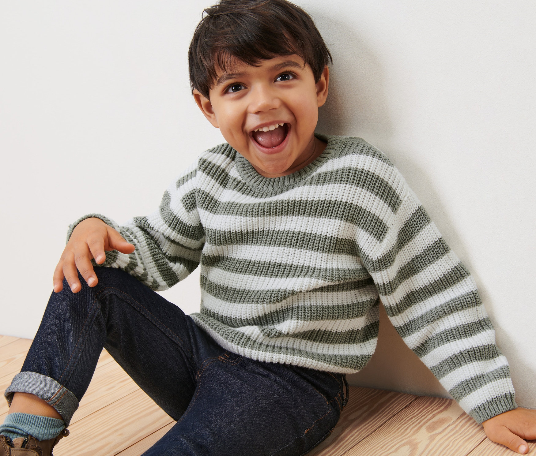 Un garçon est assis par terre et rit, il porte un pull en maille rayé pour enfant.
