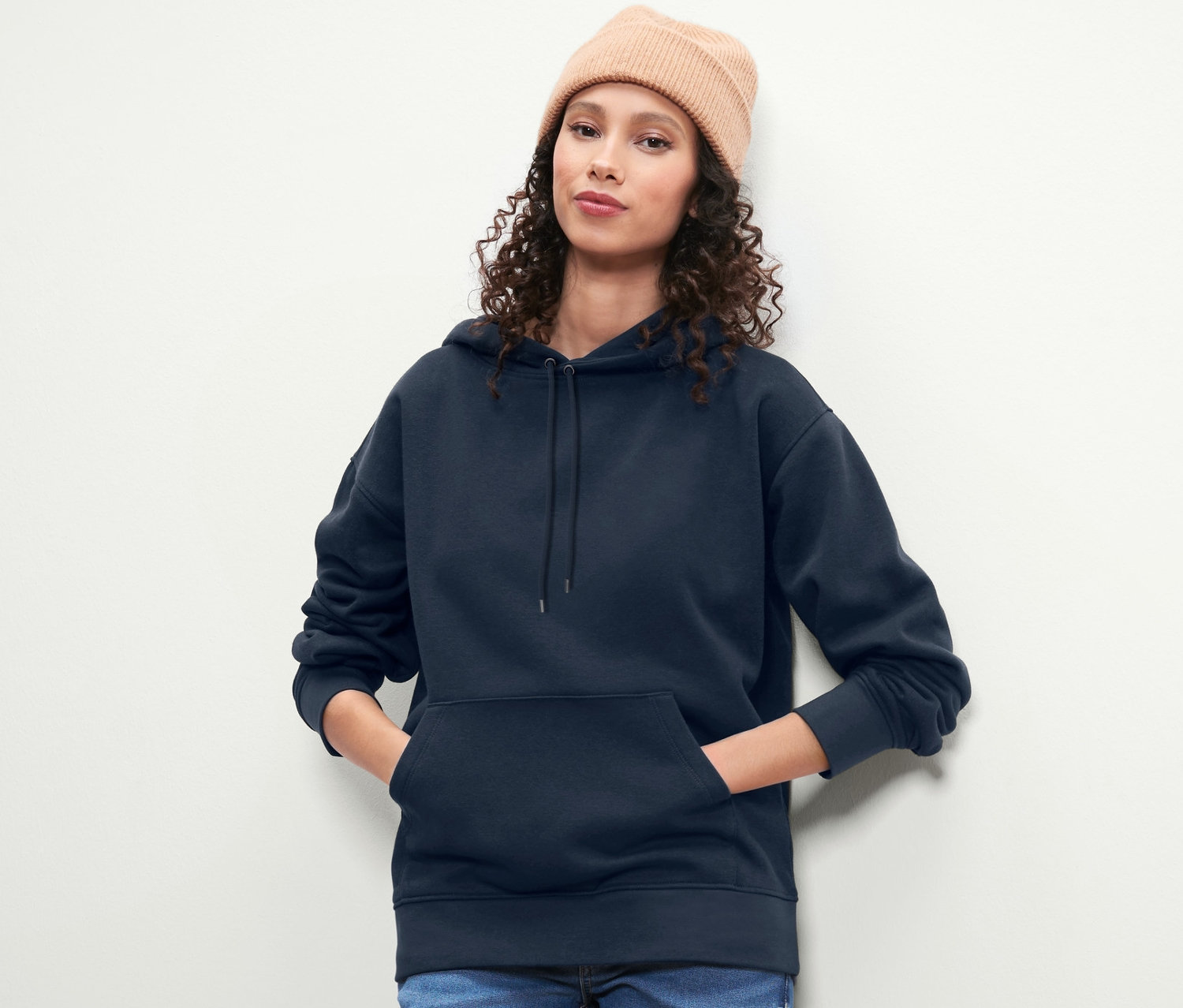 Une femme aux cheveux bouclés bruns et un bonnet pose en sweat-shirt à capuche bleu foncé.