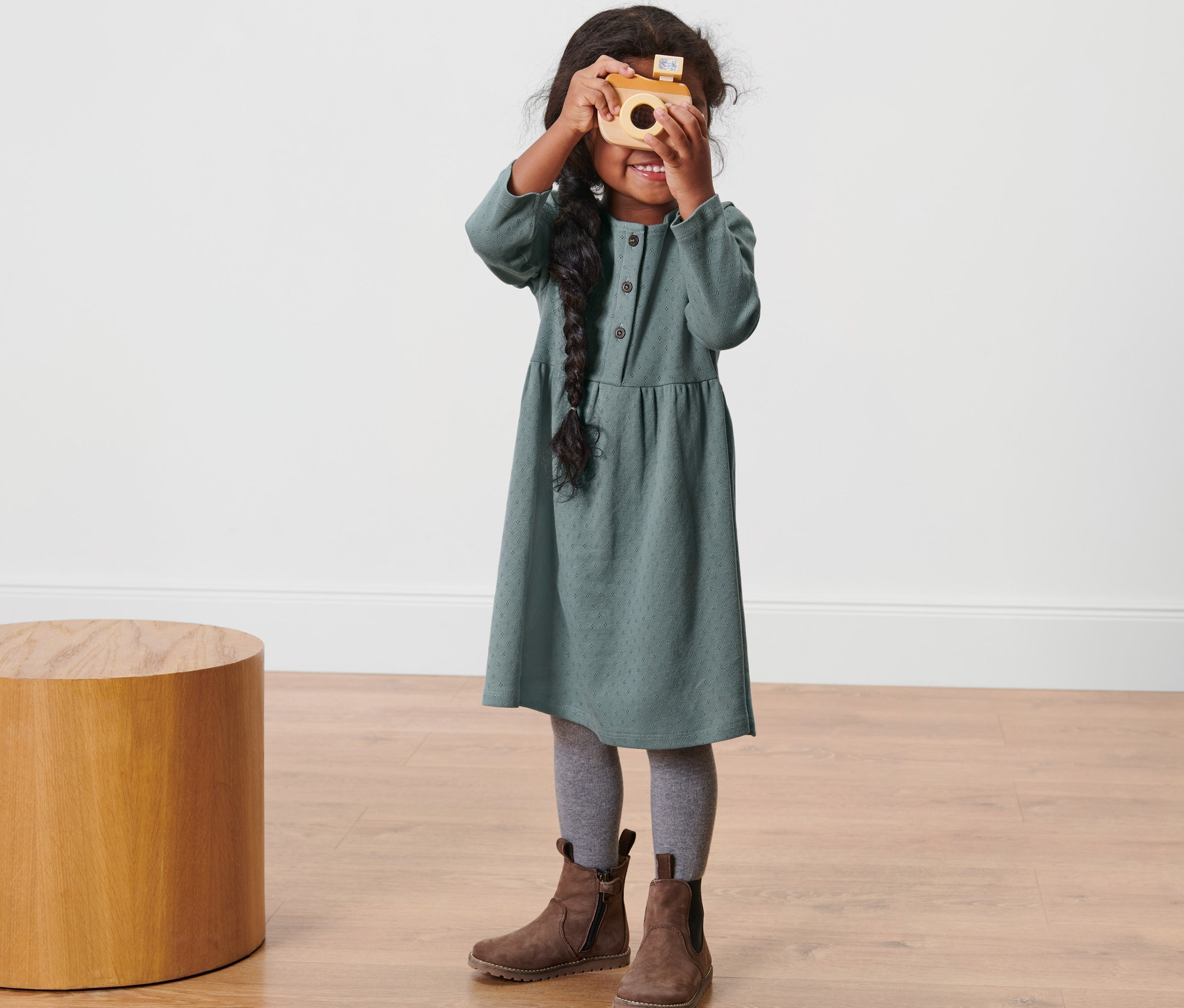 Une fillette portant une robe pour tout-petits tient un appareil photo en bois devant son visage.