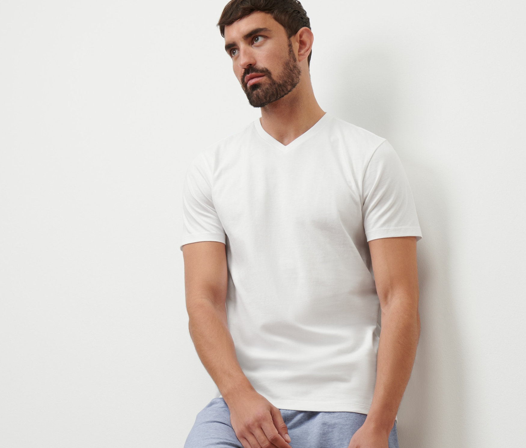 Un homme appuyé contre un mur portant un t-shirt blanc à col en V et un short bleu.