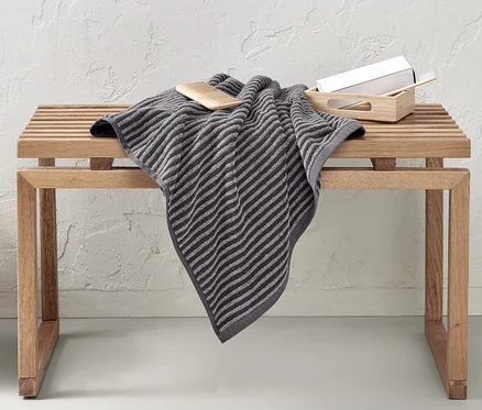 Un linge de bain de haute qualité, gris, est posé sur un banc en bois. À côté du banc se trouve un panier en osier.