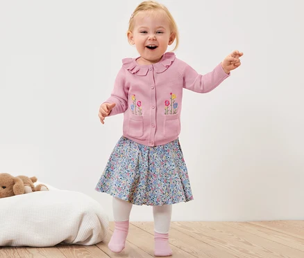 Un jeune enfant souriant agite les mains et porte une jaquette en maille rose avec des fleurs, une jupe à fleurs et des chaussettes roses.