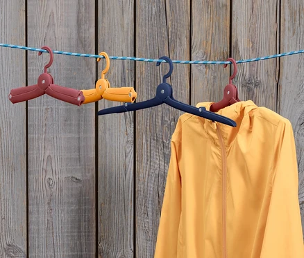 Une veste jaune est suspendue à un cintre bleu sur une corde à linge, à côté de deux 6 cintres de voyage pliables pliés : un marron et un jaune.