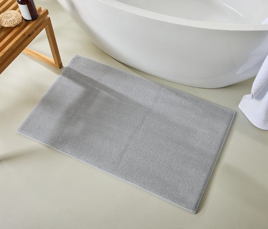 Un tapis de bain gris repose sur le sol à côté d'une baignoire. Des serviettes blanches sont posées sur un banc en bois.