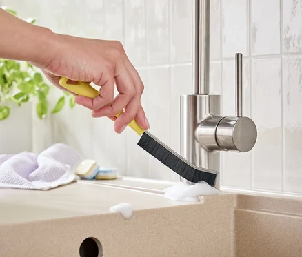 Une main nettoie un robinet avec une brosse jaune dans un évier.