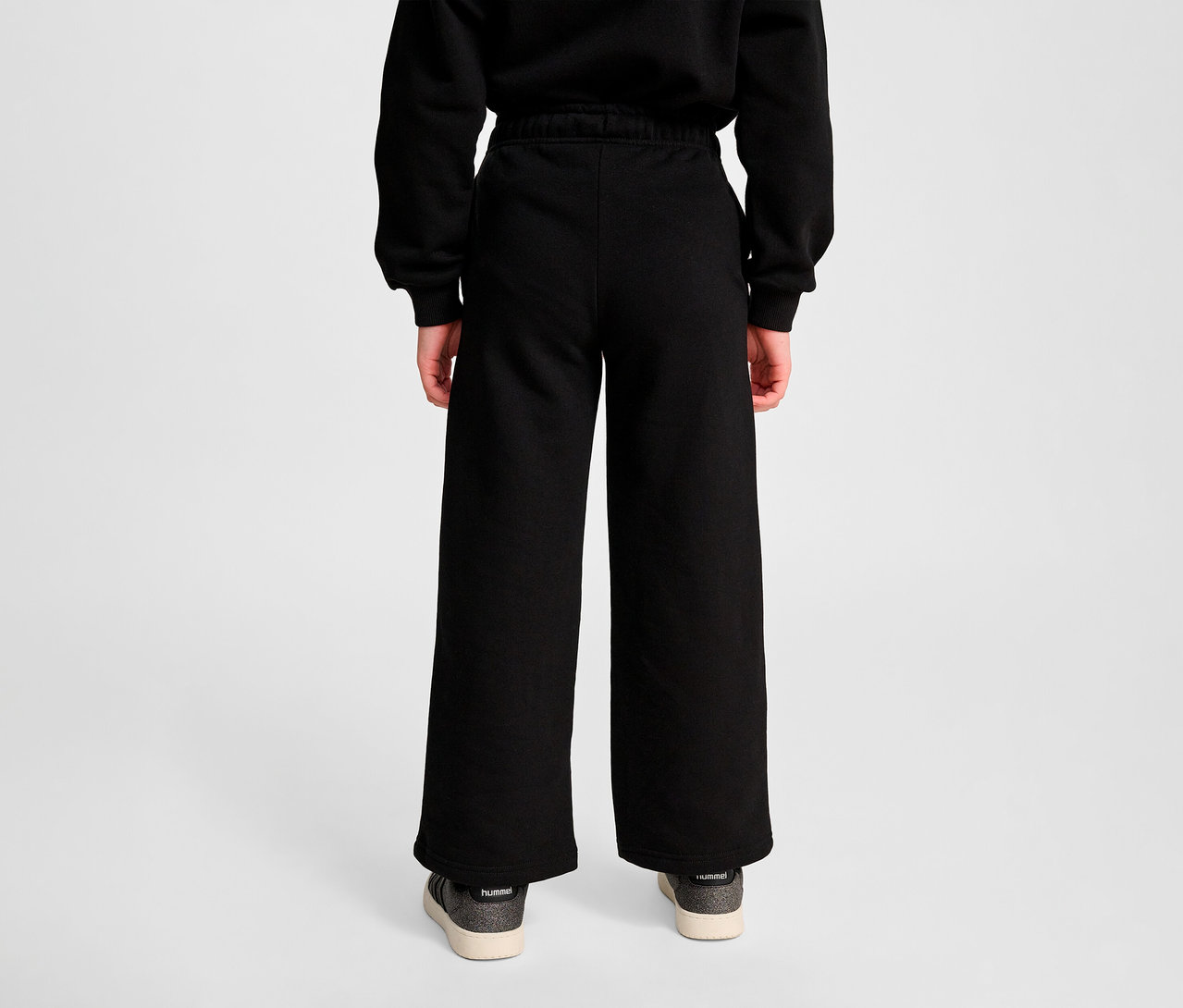 Une personne vue de dos portant un pantalon de survêtement HUMMEL HMLJR Loose WL noir et des chaussures.