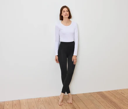 Une femme se tient devant un mur blanc, portant un t-shirt blanc à manches longues et un legging noir.