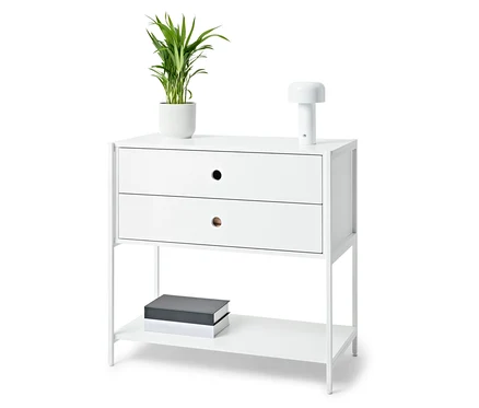 Table de chevet blanche avec deux tiroirs, une lampe, une plante en pot et des livres.