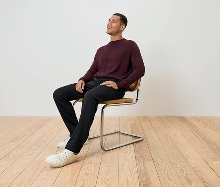 Un homme est assis sur une chaise, portant un pull bordeaux, un pantalon noir et des chaussures blanches, et regarde vers le haut.