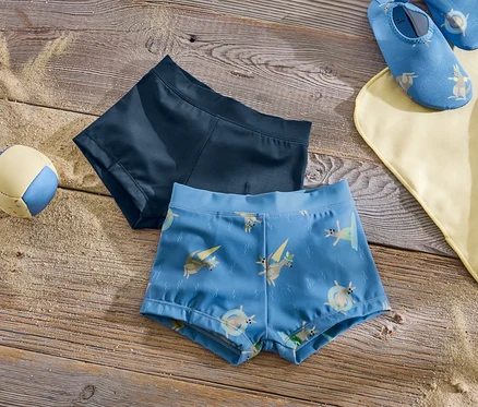 Deux maillots de bain pour enfants sont posés sur une table en bois à côté d'un ballon et d'une serviette de plage.