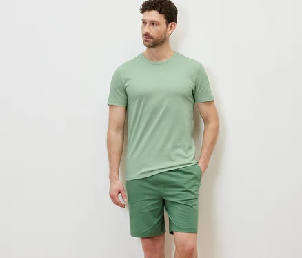 Un homme pose, portant un t-shirt vert à col rond, faisant partie d'un lot de 3, et un short vert.