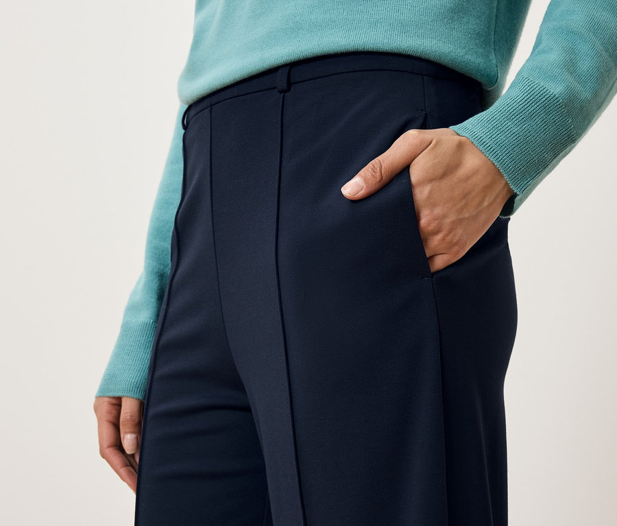 Gros plan d'une femme portant un pantalon bleu marine et un pull turquoise, une main dans la poche.
