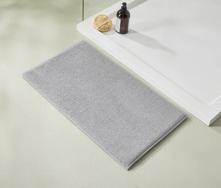 Un tapis de bain gris clair est posé à côté d'un receveur de douche blanc sur lequel reposent une bouteille marron et un luffa.