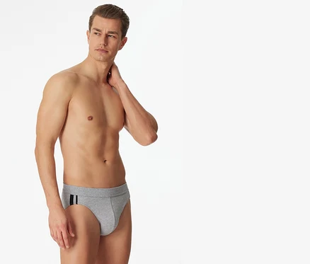 Un homme torse nu se tient debout, regardant à droite, portant un slip SCHIESSER gris chiné avec deux rayures foncées sur la hanche gauche.