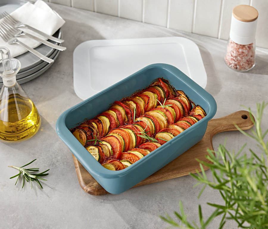 Légumes dans un plat bleu sur une planche en bois. À côté, une bouteille d'huile et un moulin à épices.