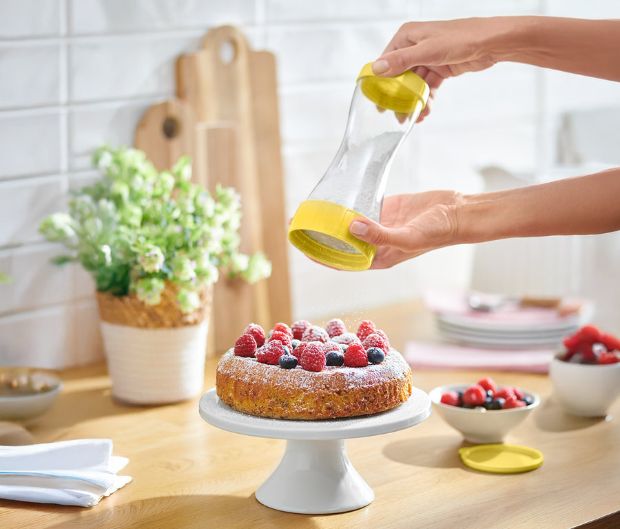 Une personne saupoudre du sucre glace sur un gâteau aux fruits rouges avec un saupoudreur jaune.